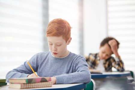 Portrait of modern red-haired schoolboy writing or taking test while sitting at desk in classroom,...