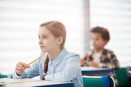 Side view portrait of cute teenage schoolgirl sitting at desk in classroom and listening to teacher, copy spaceの写真素材