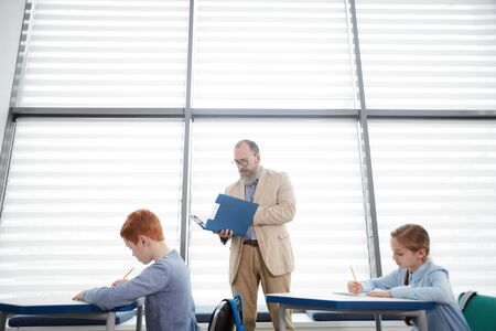 Side view at mature bearded teacher watching children taking test in school classroom, copy spaceの写真素材