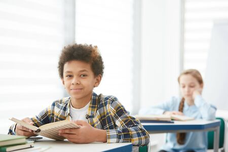 Portrait of smiling African-American boy sitting at desk in school and looking at camera while enjoying studying, copy spaceの写真素材