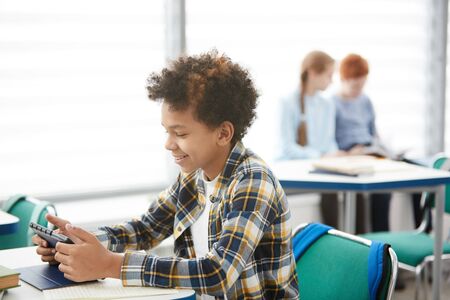 Side view portrait of smiling African-American boy using digital tablet while sitting at desk in school classroom, copy spaceの写真素材