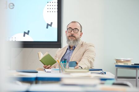 Portrait of senior bearded professor sitting at desk in school classroom and looking at camera, copy...