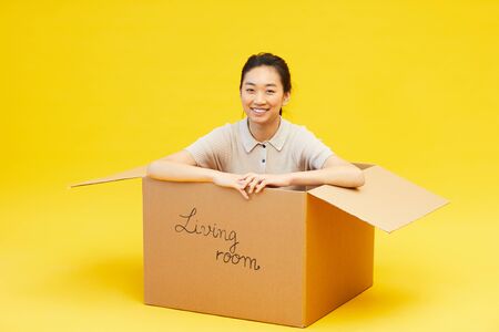 Full length portrait of young Asian woman sitting in box and smiling at camera, studio shot on...