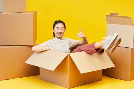 Full length portrait of happy Asian woman sitting in box and showing thumbs up, studio shot on...