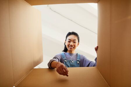 Low angle view at smiling Asian woman reaching into cardboard box while packing or unpacking for new...