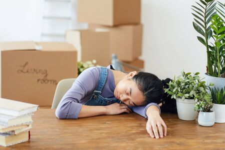 Portrait of exhausted Asian woman sleeping on wooden table while packing for moving or relocation to...