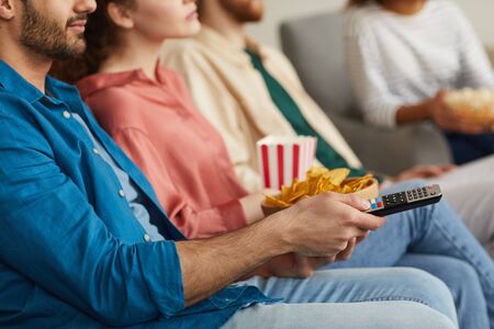 Close up of multi-ethnic group of friends watching TV together while sitting on cozy sofa at home...