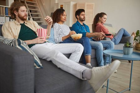 Low angle view at multi-ethnic group of friends watching TV together while sitting on comfortable...