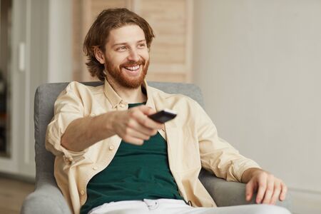 Portrait of modern bearded man watching TV at home while relaxing in comfy armchair and holding remote, copy spaceの写真素材