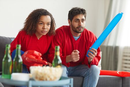 Portrait of modern mixed-race couple watching sports on TV at home and cheering for intense match...
