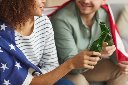 Close up of mixed-race young couple clinking beer bottles while wearing American flag, copy spaceの写真素材