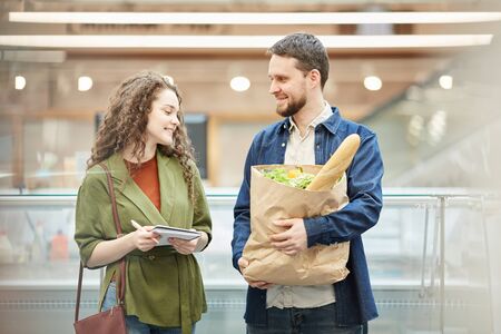 Waist up portrait of cheerful couple holding paper bag with groceries while enjoying shopping in supermarket, copy spaceの写真素材
