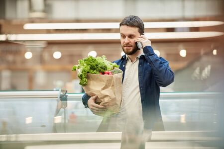 Waist up portrait of adult bearded man calling by smartphone while grocery shopping in supermarket,...