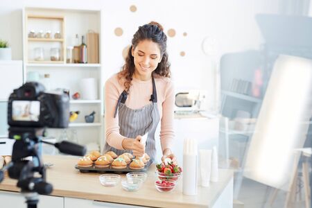 Professional baker standing at home kitchen decorating fresh cupcakes with sweet cream, shooting...