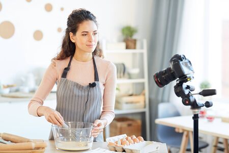 Good-looking young woman wearing apron standing at table in modern kitchen beating up eggs on camera for her food blogの写真素材