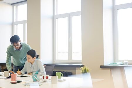 Horizontal shot of young African American man helping his female colleague with work in spacious office room, copy spaceの写真素材