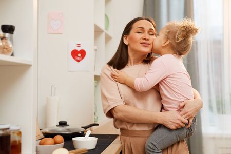 Cute little child tenderly kissing her happy young mother while sitting in her arms, horizontal medium shotの写真素材