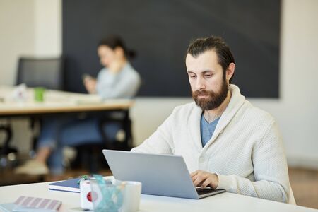 Handsome young man with beard on face sitting at office table working on his laptop with serious facial expressionの写真素材