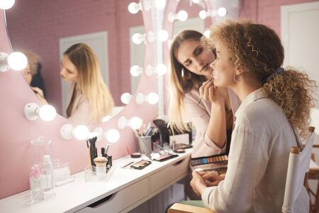 Side view portrait of female makeup artist styling fashion model by vanity mirror in pink dressing room interior, copy spaceの写真素材