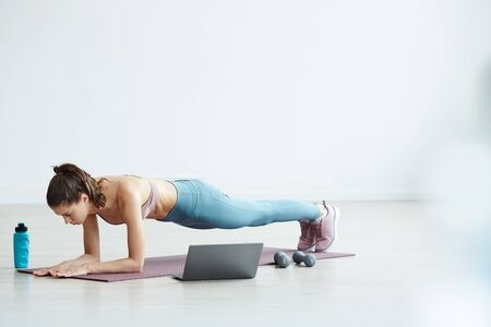 Minimal full length portrait of fit young woman doing plank exercise while watching workout video...