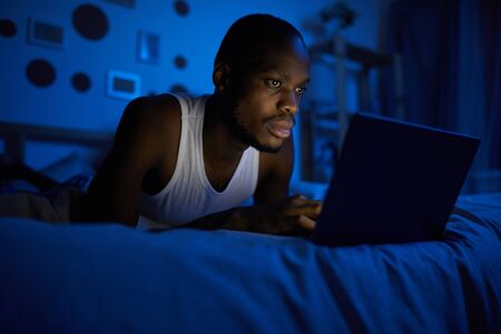 Portrait of young African-American man using laptop while lying on bed at night, copy spaceの写真素材