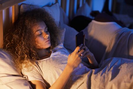 High angle portrait of curly-haired young woman using smartphone in bed at night while lying next to...