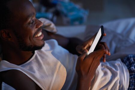 High angle portrait of smiling African-American man using smartphone in bed at night, copy spaceの写真素材