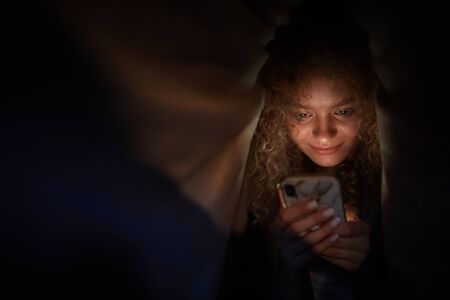 Portrait of curly haired young woman looking at smartphone screen and smiling while lying in bed in...