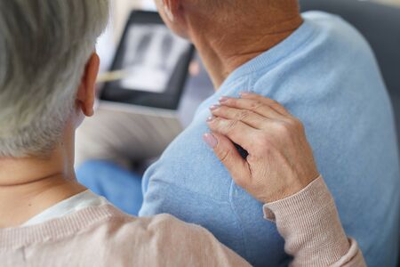 Close up of senior woman supporting husband during visit to medical clinic, focus on female hand on shoulder, copy spaceの写真素材