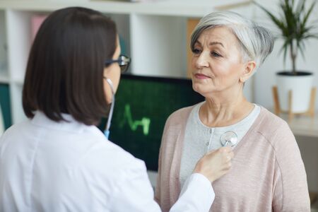 Portrait of female doctor listening to heart and breathing of senior woman via stethoscope on chest during consultation in clinicの写真素材