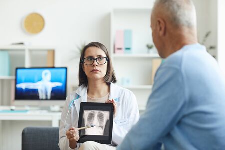 Portrait of serious female doctor holding x-ray image of lungs and chest while consulting senior...