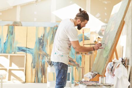 Side view portrait of contemporary bearded artist painting picture on easel while working in spacious art studio lit by sunlight, copy spaceの写真素材
