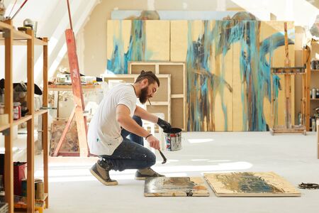 Side view full length portrait of contemporary bearded artist painting abstract picture while sitting on floor in art studio lit by sunlight, copy spaceの写真素材
