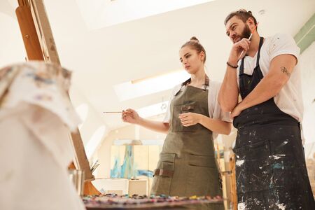 Warm-toned low angle view at creative couple painting picture together while standing by easel in...