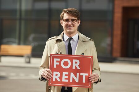 Waist up portrait of male real estate agent smiling happily looking at camera while holding red FOR RENT sign outdoors, copy spaceの写真素材