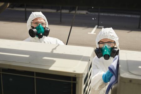 High angle view at two workers wearing hazmat suits and respirators disinfecting surfaces outdoors lit by sunlight, copy spaceの写真素材