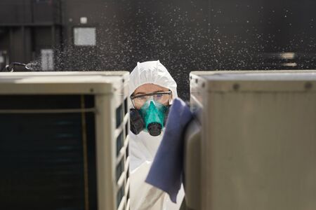 Portrait of female worker wearing protective suit and respirator disinfecting surfaces outdoors lit by sunlight, copy spaceの写真素材