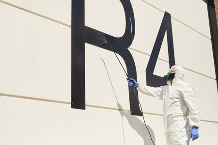Wide angle view at worker wearing protective gear and spraying chemicals over black building facade...