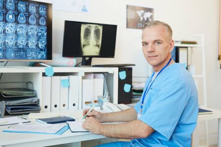 Portrait of handsome mature doctor looking at camera while posing at workplace in modern clinic interior with CT scans in background, copy spaceの写真素材