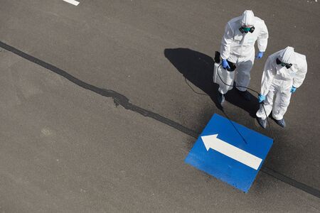 Above view at two workers wearing protective gear and spraying chemicals outdoors standing on concrete road with blue arrow pointing left, copy spaceの写真素材