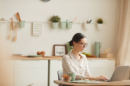 Warm-toned side view portrait of elegant young woman wearing glasses while using laptop at cozy home office workplace, copy spaceの写真素材