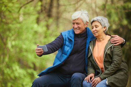 Portrait of active senior couple taking selfie photo via smartphone while resting during hike in autumn forest, copy spaceの写真素材