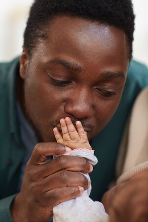 Vertical close up portrait of young African-American father kissing tiny hand of unrecognizable mixed-race babyの写真素材