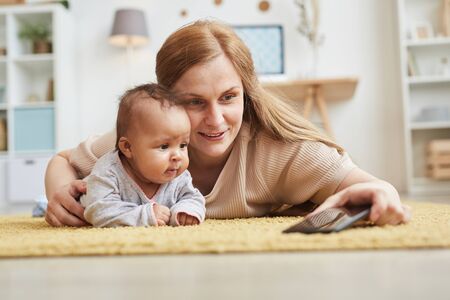 Portrait of mature mother taking selfie photo with cute mixed race baby while playing on carpet at...