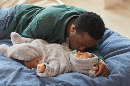 Portrait of loving African-American father kissing cute baby while lying on bed in cozy home...