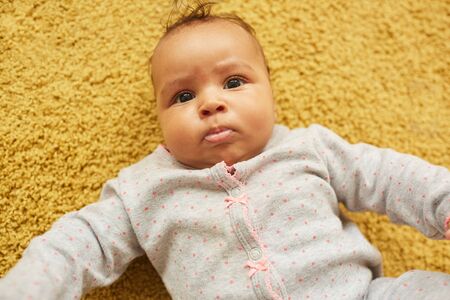 Above view portrait of cute mixed-race baby looking at camera while lying on fluffy yellow carpet,...