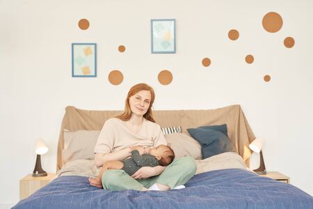 Warm-toned full length portrait of happy adult mother breastfeeding mixed-race baby and looking at camera while sitting on bed at home, copy spaceの写真素材