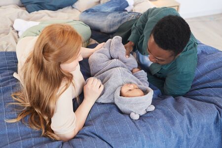 Portrait of modern interracial family playing with cute mixed-race baby while lying on comfortable...