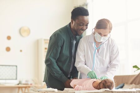 Waist up portrait of female doctor examining baby with smiling African-American father standing by them, copy spaceの写真素材