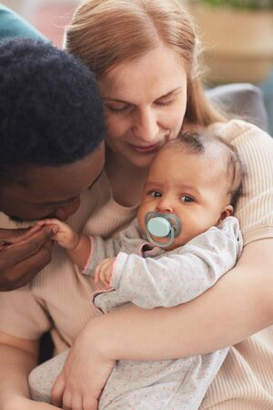 Vertical warm-toned portrait of loving interracial family holding baby boy, with African-American...